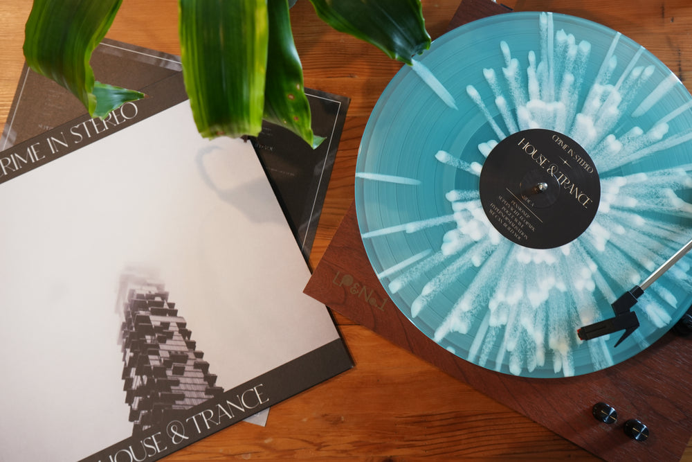 A blue and white marbled Electric Blue Vinyl LP by Crime In Stereo spins on the turntable. A Presale Album cover with a blurry tower and another sleeve rest under a green plant leaf nearby—ideal for House & Trance vinyl lovers.