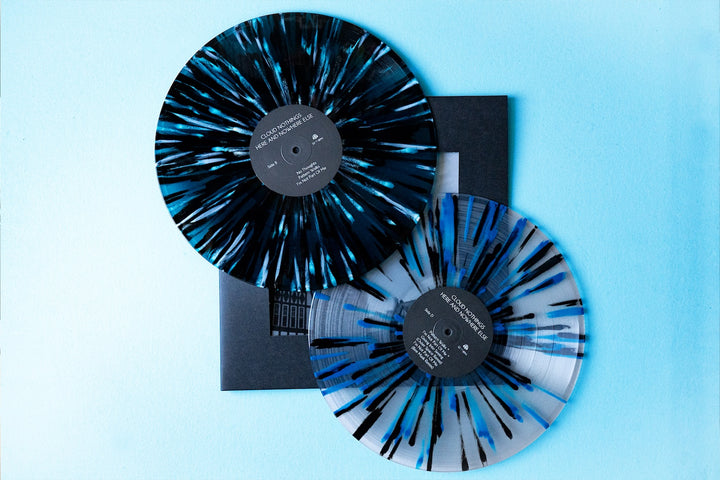 Two blue and black splatter-patterned vinyl records from Cloud Nothings’ Live at Bells Brewery presale are displayed on a light blue surface, slightly overlapping atop a dark record sleeve.