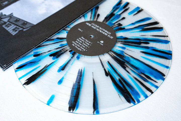 A white vinyl record with black and blue splatter, featuring “Live at Bells Brewery” by Cloud Nothings, rests partly in a black sleeve. The center label displays track names, album info, and presale details.