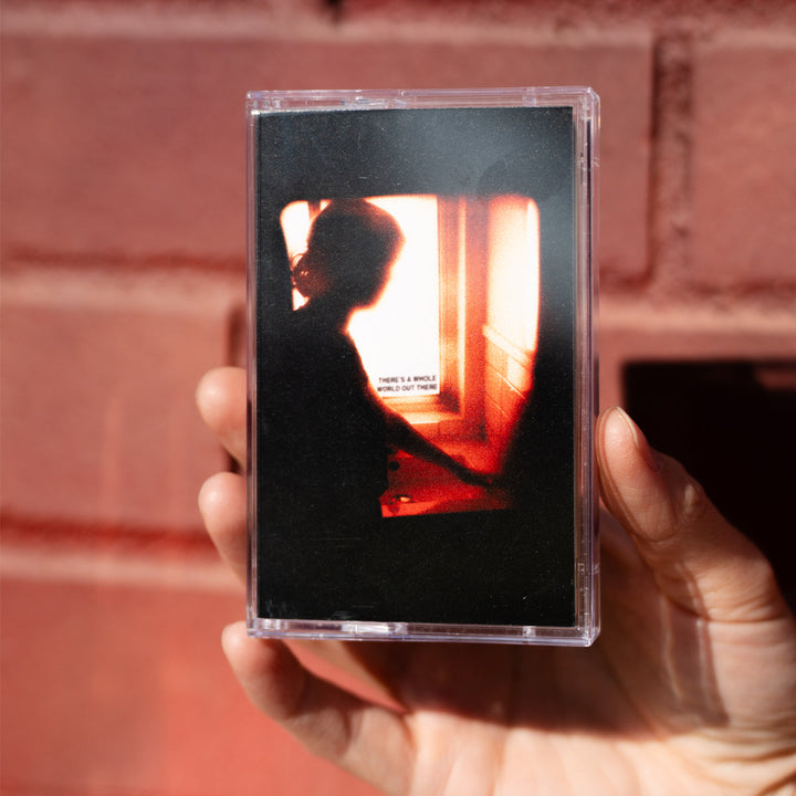 A hand holds an Arm’s Length cassette tape with cover art showing a person’s silhouette by a sunlit window and small text, “THERES A WHOLE WORLD OUT THERE”—perfect for fans awaiting the album release.