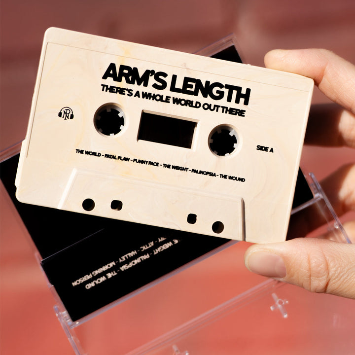 A hand holds a beige cassette labeled Arms Length: There’s a Whole World Out There, with tracklist below. The presale tape case sits open on a blurred pink background.