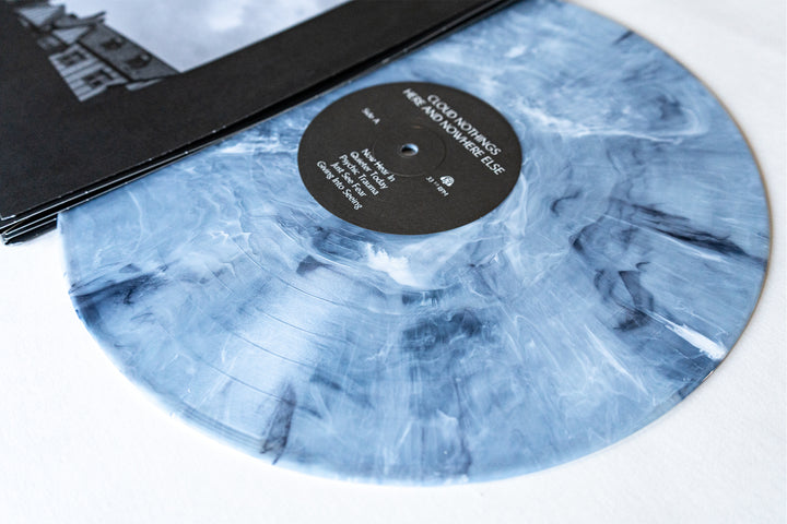 A blue and white marbled Cloud Nothings vinyl record peeks from its black sleeve, the label reads Here and Nowhere Else. This presale item promises an exciting tracklist as it rests on a white surface.