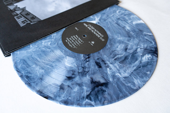 A close-up of a marbled blue and white vinyl record by Cloud Nothings, partially out of its sleeve, reveals the label with the album title Here and Nowhere Else and its track list.