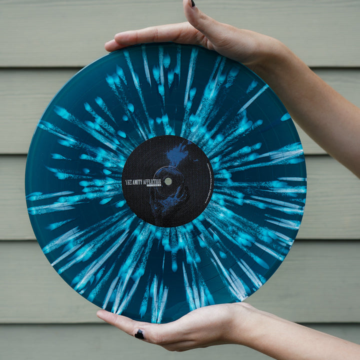 A person with black nail polish holds a Royal Blue Vinyl splatter record by The Amity Affliction in front of a tan paneled wall, hinting at the exclusive Redux Tracklist for the September 2024 release.