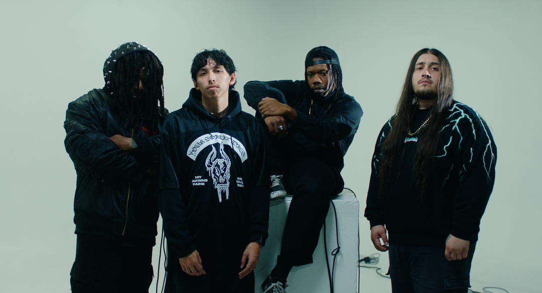 Four men in black outfits stand in front of a brick wall, posing confidently. One points at the camera, while the others stand with relaxed postures. All are wearing casual shoes.