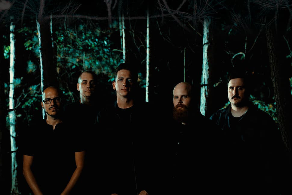 Five men stand in a forest, looking into the camera. The photo is black and white, with light shining through the trees and a vignette effect around the edges.
