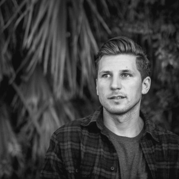 Black and white photo of a young man with short hair, wearing a plaid shirt over a t-shirt, looking slightly to the side. Blurred foliage is in the background.