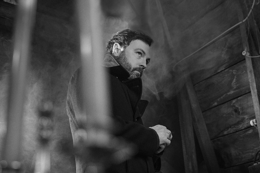 A man with a beard and coat stands in a dimly lit, smoky wooden room, looking to the side with a serious expression.