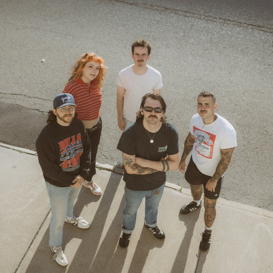 Five people stand outside on a sidewalk, looking up at the camera. Three men and two women wear casual clothes, including jeans, shorts, t-shirts, and sneakers, with bright sunlight casting shadows.