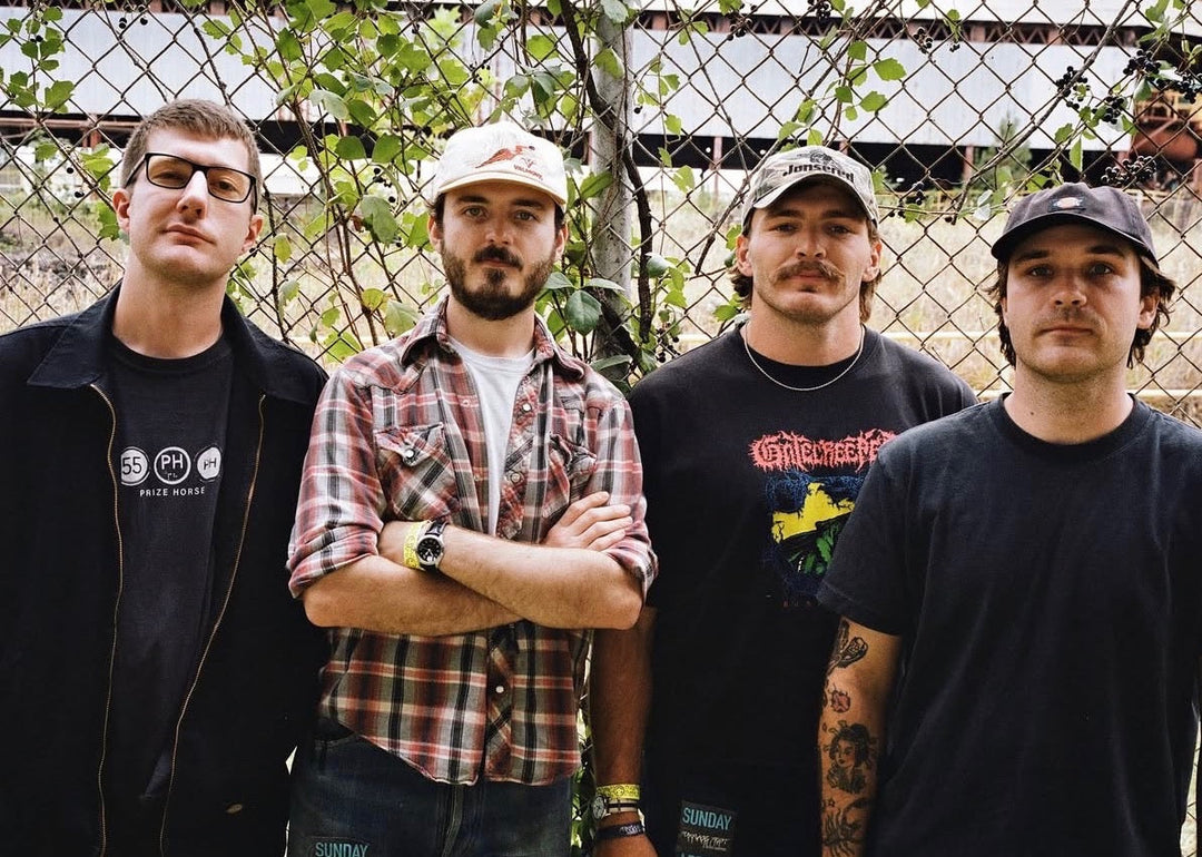 Four men stand in front of a chain-link fence covered in vines. They wear casual clothes—T-shirts, a plaid shirt, and caps—and look directly at the camera with neutral expressions.