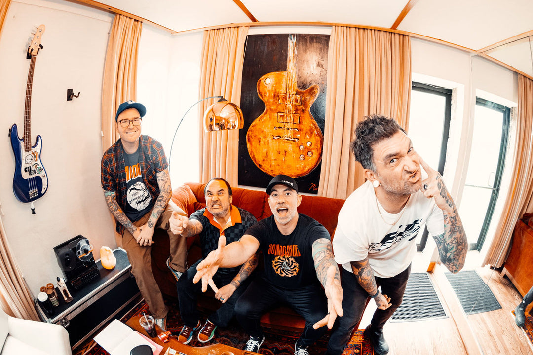 Four men with tattoos enthusiastically pose in a room with a hanging guitar decoration, music equipment, and a bass guitar on the wall, giving energetic and playful expressions.