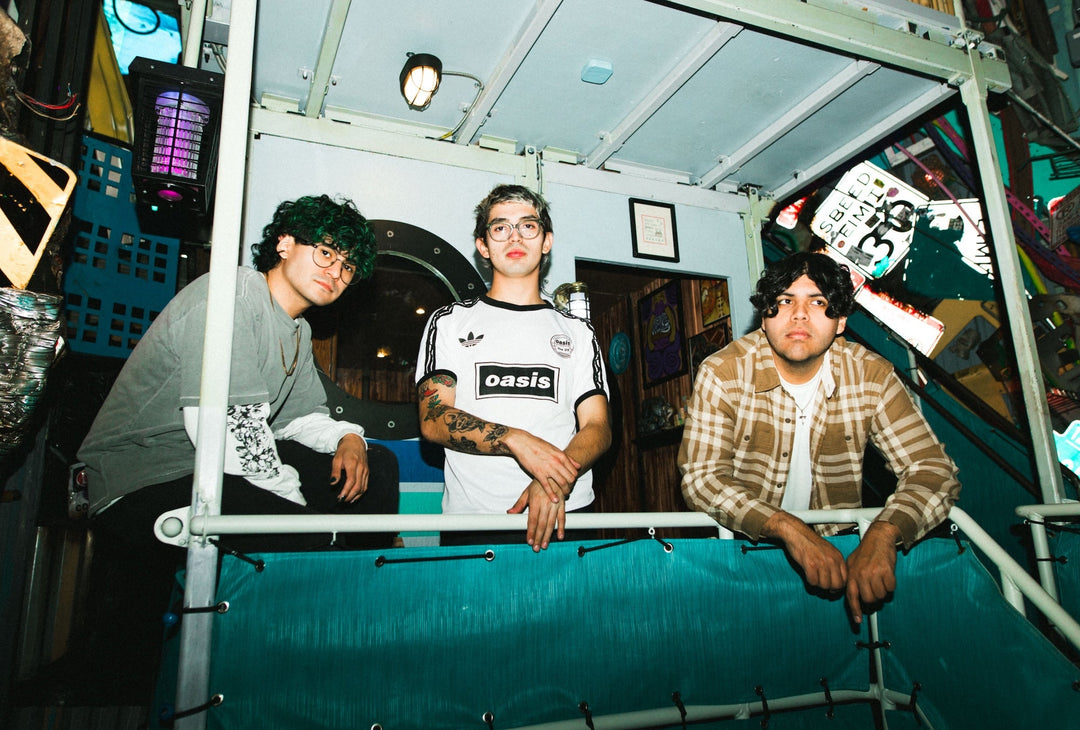 Three young men pose indoors on a metal staircase, surrounded by colorful, eclectic decorations and neon lights. Two wear glasses, and one has visible tattoos and an Oasis soccer jersey.