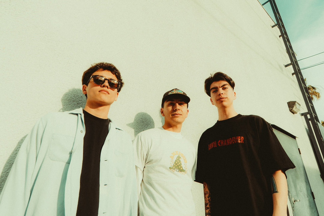 Three young men stand outside against a light-colored wall, looking at the camera. One wears sunglasses, another a cap, and all have casual clothing. The sky is blue in the background.