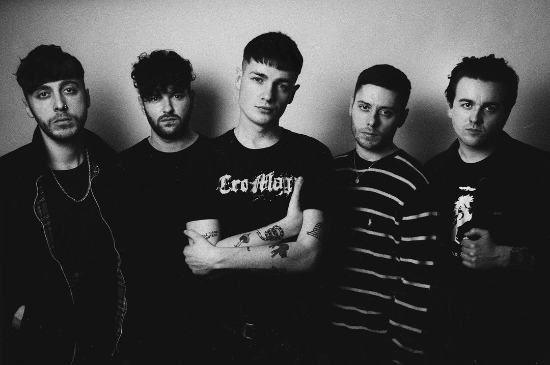 Five young men stand close together against a plain background, looking seriously at the camera. The photo is in black and white, with high-contrast lighting and a moody atmosphere.