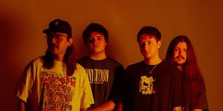 Four young men stand side by side against an orange background, all wearing casual dark shirts and looking in different directions with neutral expressions.