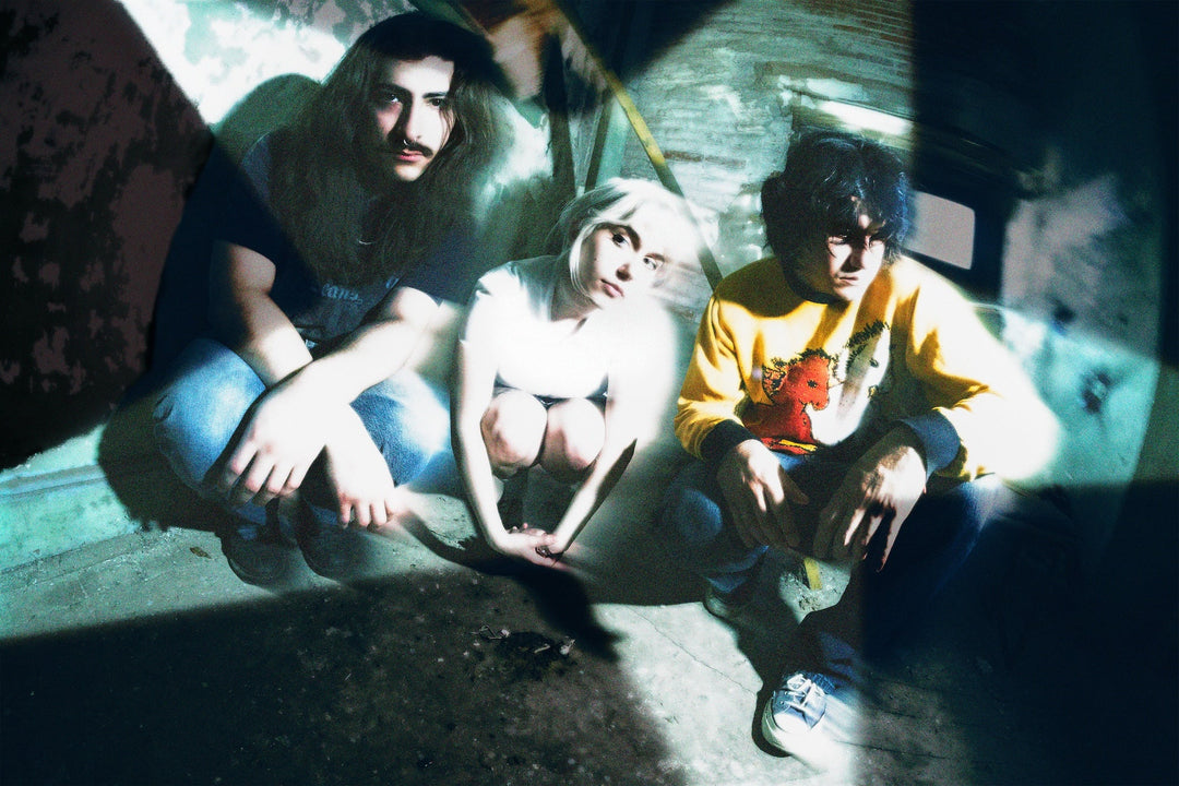 Three young adults sit on the floor in a dim, grungy room with dramatic lighting and shadows. The image has a slightly distorted, artistic effect.