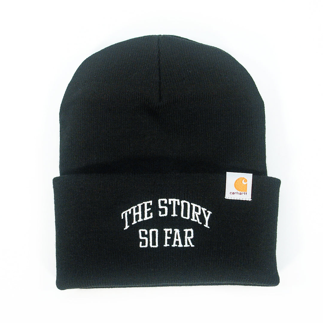 A black winter beanie by The Story So Far with a folded brim, white embroidered "THE STORY SO FAR" text on the front, and a small Carhartt logo tag on the side.