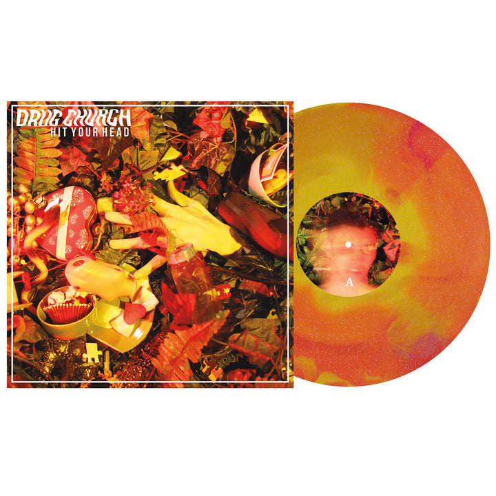 The image displays Drug Church's Hit Your Head vinyl. Its cover has scattered yellow plastic hands, party favors, and red-yellow leaves; the yellow-orange Glitter Vinyl LP features a blurred face at the center.