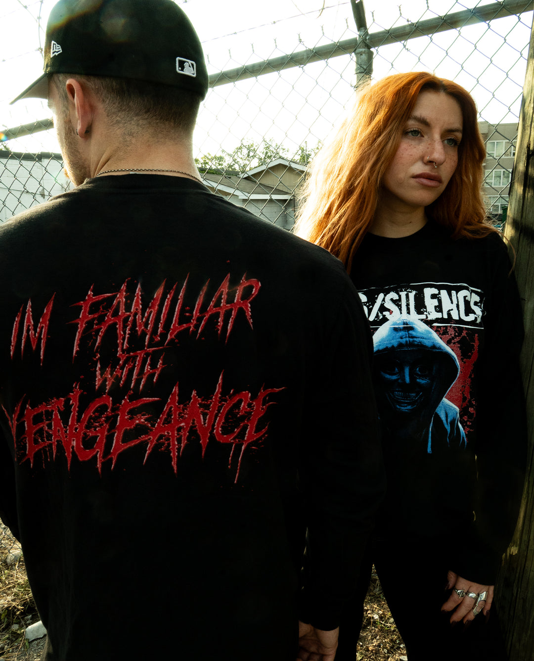 Two people stand near a chain-link fence. The man faces away, wearing a shirt that says IM FAMILIAR WITH VENGEANCE. The woman, with long red hair, faces the camera, wearing a black Comfort Colors long sleeve crafted from soft ring spun cotton and finished with a bold graphic design.