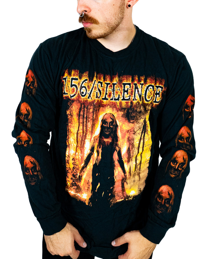 A person wearing a black Comfort Colors long sleeve shirt with 156/SILENCE and a horror-themed graphic of a figure in flames. Made from soft ring spun cotton and garment dyed for lasting quality, the sleeves feature multiple red skulls. The person is looking to the side.