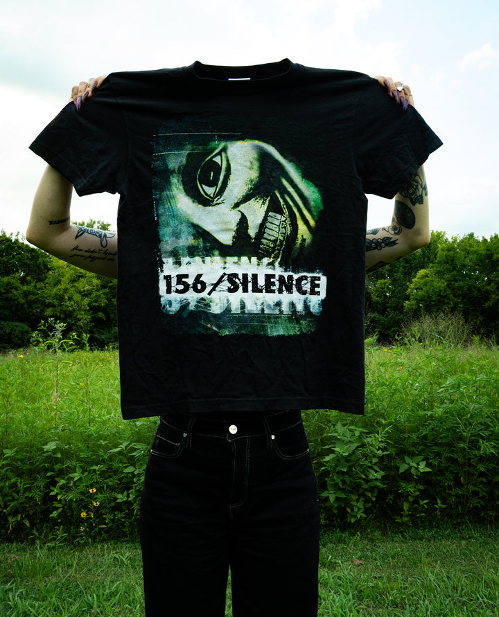 Person holding up a black Comfort Colors T-Shirt with a graphic of a fish head and the text 156/SILENCE, standing outdoors in front of green bushes and trees under a cloudy sky.