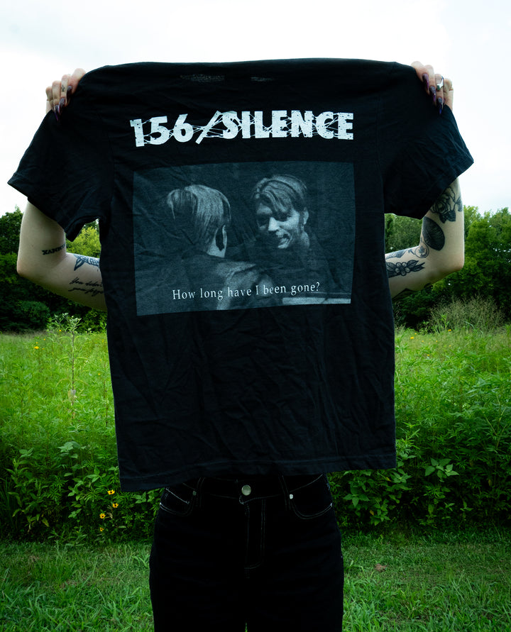 A person holds up a black Comfort Colors T-Shirt featuring 156/Silence and a black-and-white image of two people. Text on the shirt reads, How long have I been gone? Green bushes and trees are in the background.