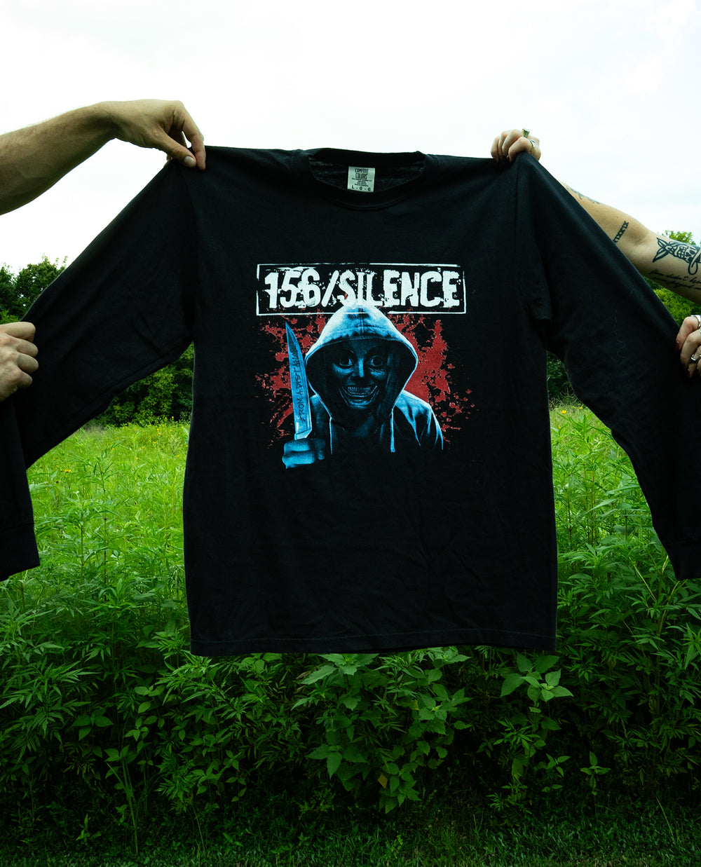 Two people hold up a black Comfort Colors long sleeve shirt outdoors. The garment dyed shirt features a hooded figure with a knife, red splatter background, and the text 156/SILENCE above the image. Green plants and sky fill the background, highlighting the ring spun cotton’s soft texture.