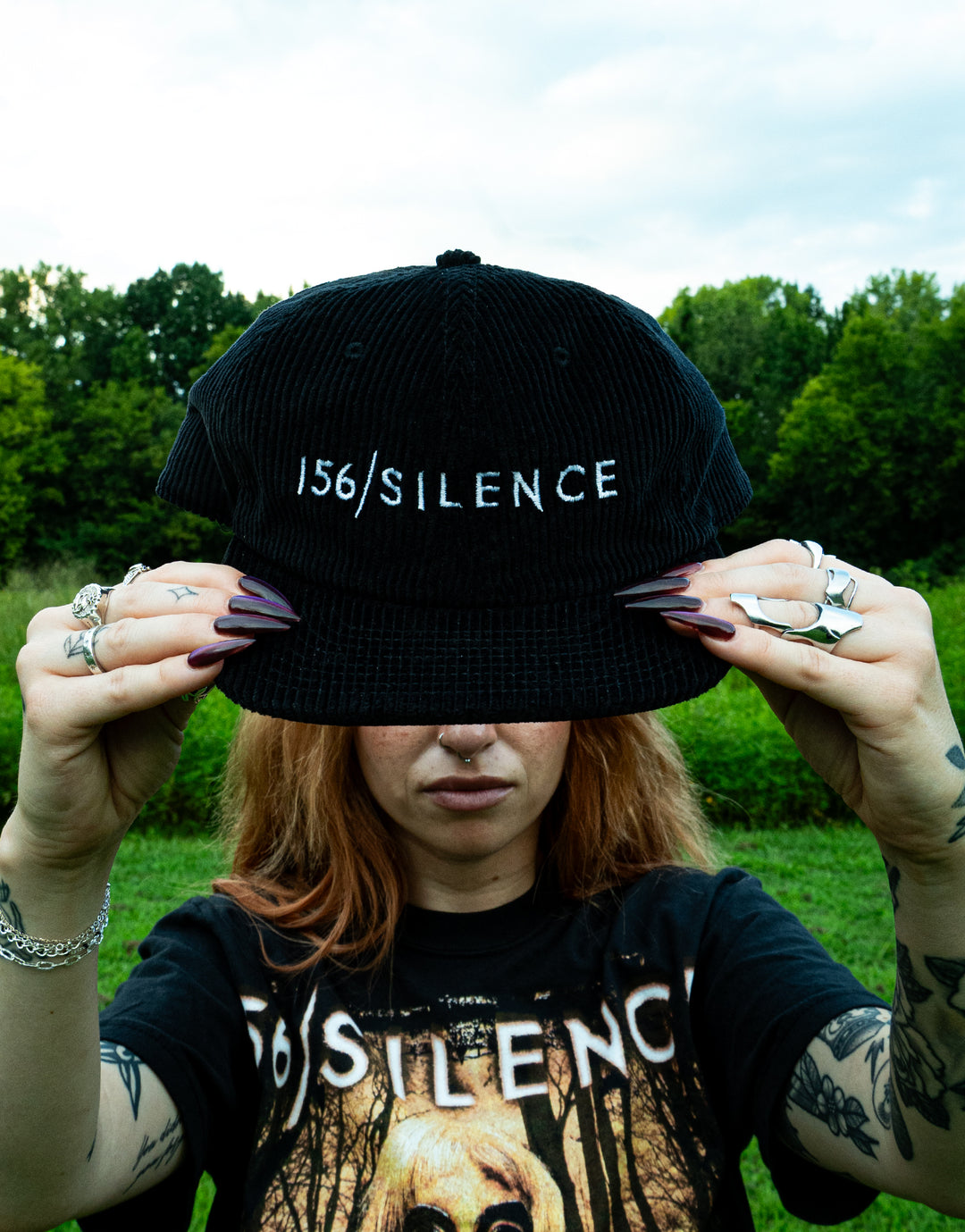A person with long hair holds a black embroidered cord cap in front of their face, showing the text 156/SILENCE. They wear rings, have dark nails, and are dressed in a matching 156/SILENCE shirt by AS Colour. Green trees and grass are in the background.