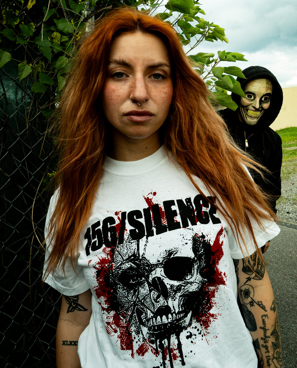 A woman with long red hair and tattoos wears a 156/SILENCE skull graphic shirt made from soft, heavyweight fleece, standing outdoors. Behind her, a person in a hooded jacket and skull mask stares at the camera through green foliage.