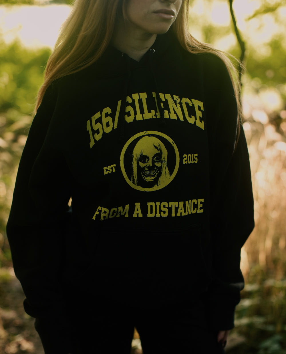 A person stands outdoors wearing a black, heavyweight pullover hoodie with yellow text reading “156/Silence Est 2015 From a Distance” and an illustration of a creepy face. The cozy fleece lined hood and premium ring-spun cotton fabric add comfort and durability to this striking design.