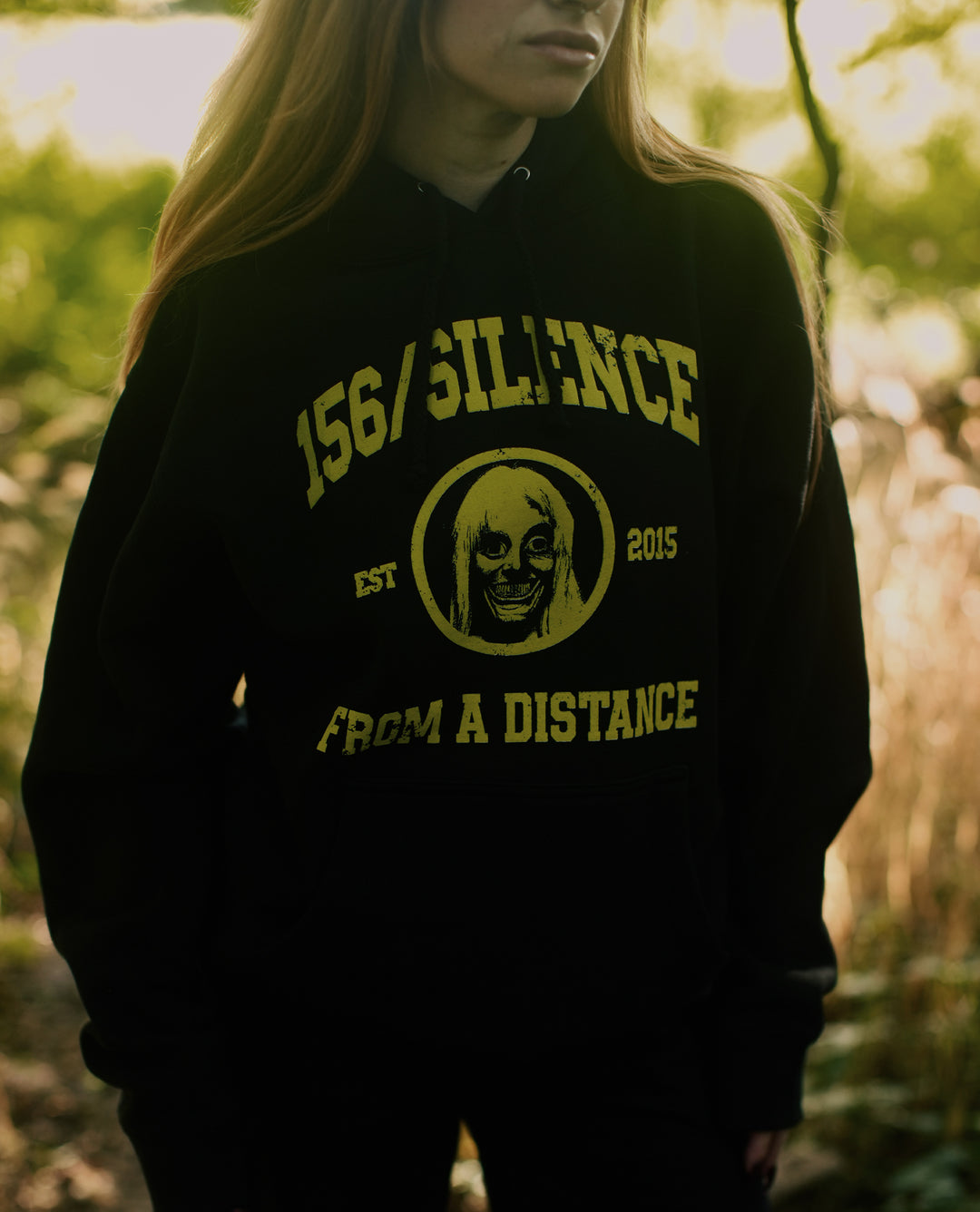 A person stands outdoors wearing a black, heavyweight pullover hoodie with yellow text reading “156/Silence Est 2015 From a Distance” and an illustration of a creepy face. The cozy fleece lined hood and premium ring-spun cotton fabric add comfort and durability to this striking design.