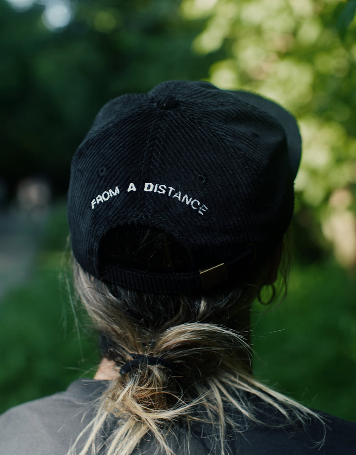 Person with light brown hair in a low ponytail wearing a black AS Colour cord cap that reads FROM A DISTANCE in white embroidered letters, standing outdoors with greenery in the background.