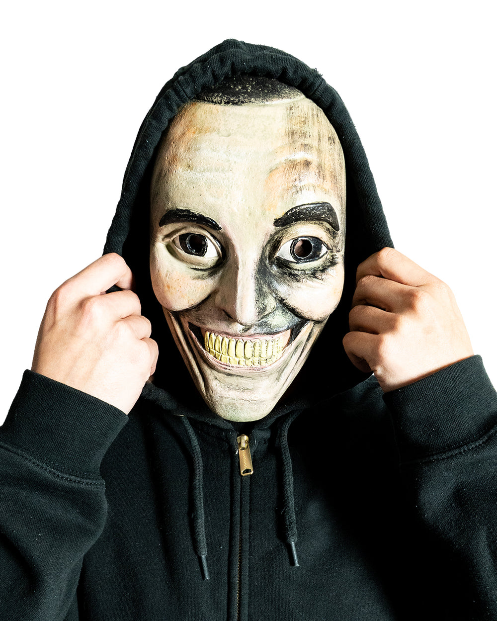A person in a black hoodie holds the hood up, wearing a creepy, exaggerated smiling mask with big eyes and yellow teeth, against a white background.