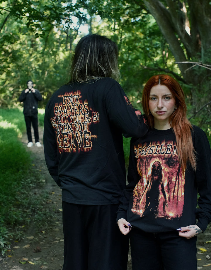 Two people in black horror-themed shirts—crafted from soft ring spun cotton—stand on a wooded path, one facing the camera and one with their back to it. A third person in dark clothes and a white mask stands further down the path.