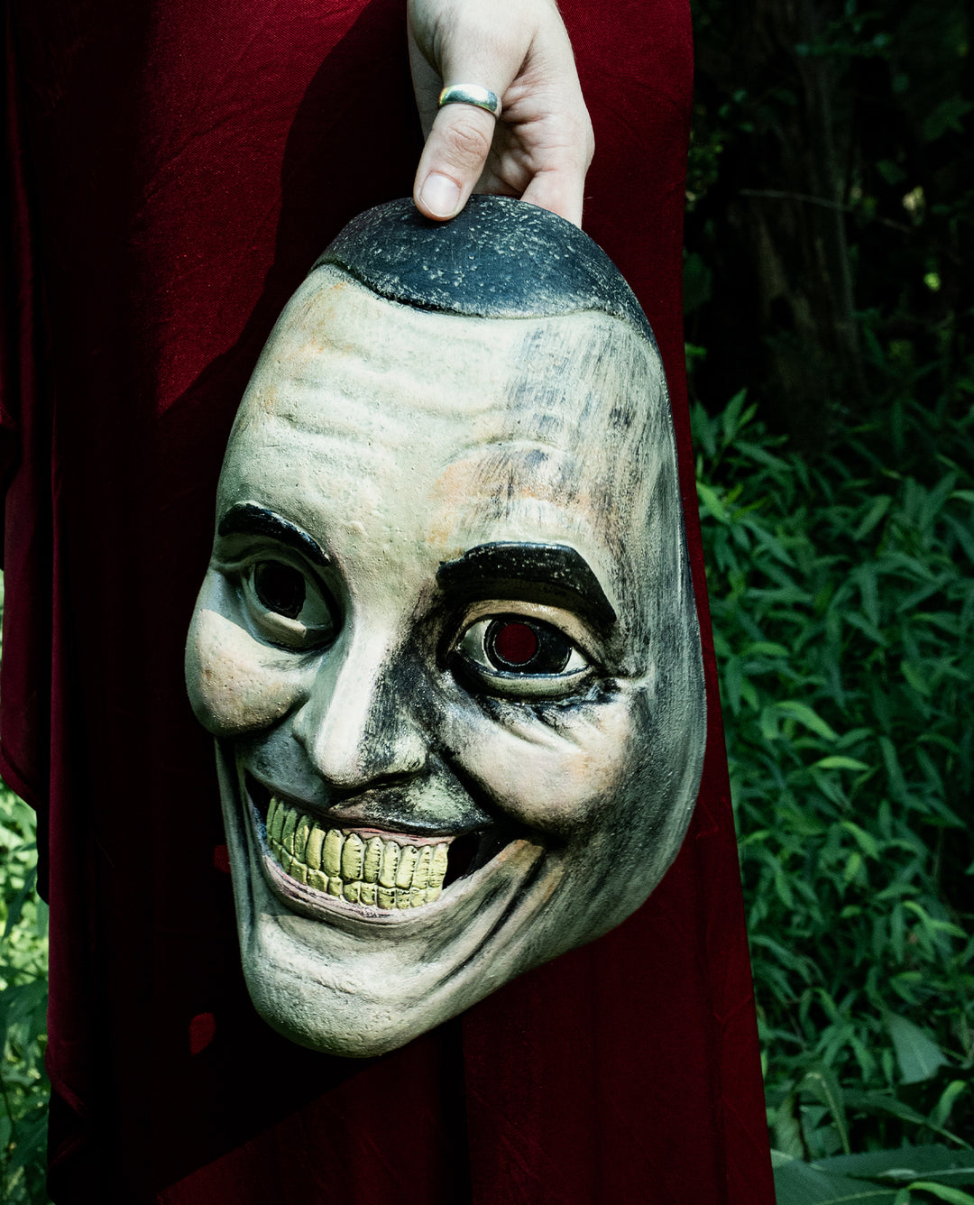 A person in a red garment holds a creepy, grinning mask with yellow teeth and dark, arched eyebrows, standing against a background of green foliage.