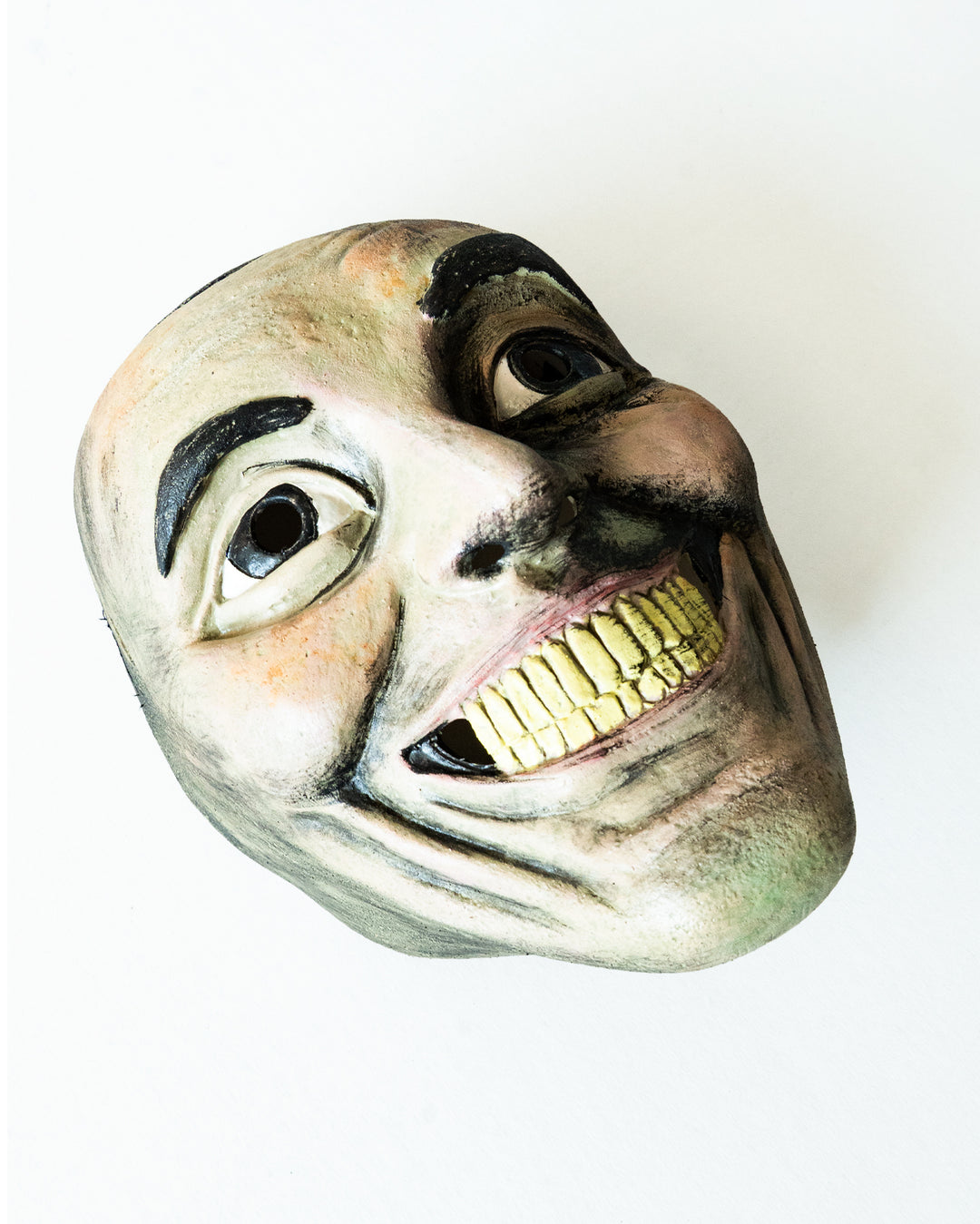 A close-up of a creepy, exaggerated smiling mask with yellow teeth and dark eye holes, lying on a white surface.