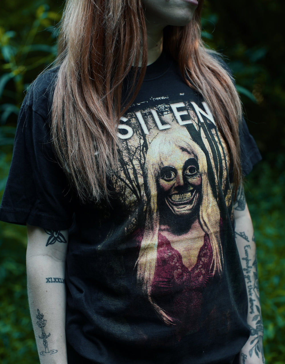A person with long, light brown hair wears a black Comfort Colors T-Shirt featuring eerie art of a wide-eyed, grinning woman in the woods. Their tattooed arms are visible, and the background is lush greenery. The garment dyed, ring-spun cotton adds both comfort and character to their unique style.