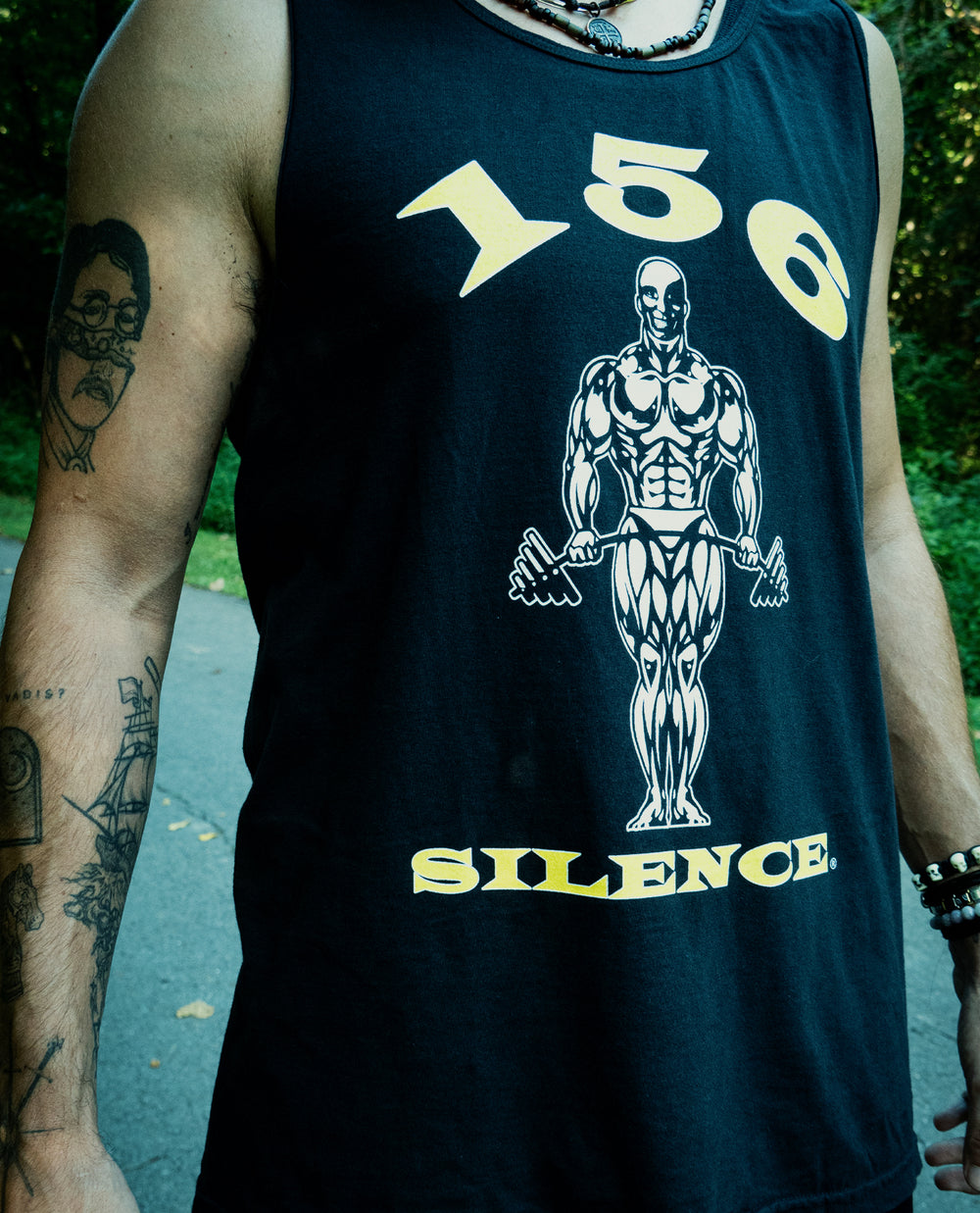 A person wearing a black Comfort Colors tank top featuring a muscular figure, the numbers 150, and the word SILENCE. Visible tattoos are on their arm, and they are standing outdoors on a path.