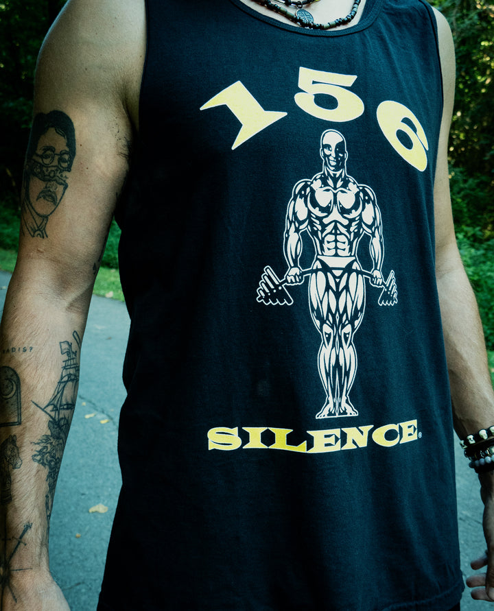A person wearing a black Comfort Colors tank top featuring a muscular figure, the numbers 150, and the word SILENCE. Visible tattoos are on their arm, and they are standing outdoors on a path.