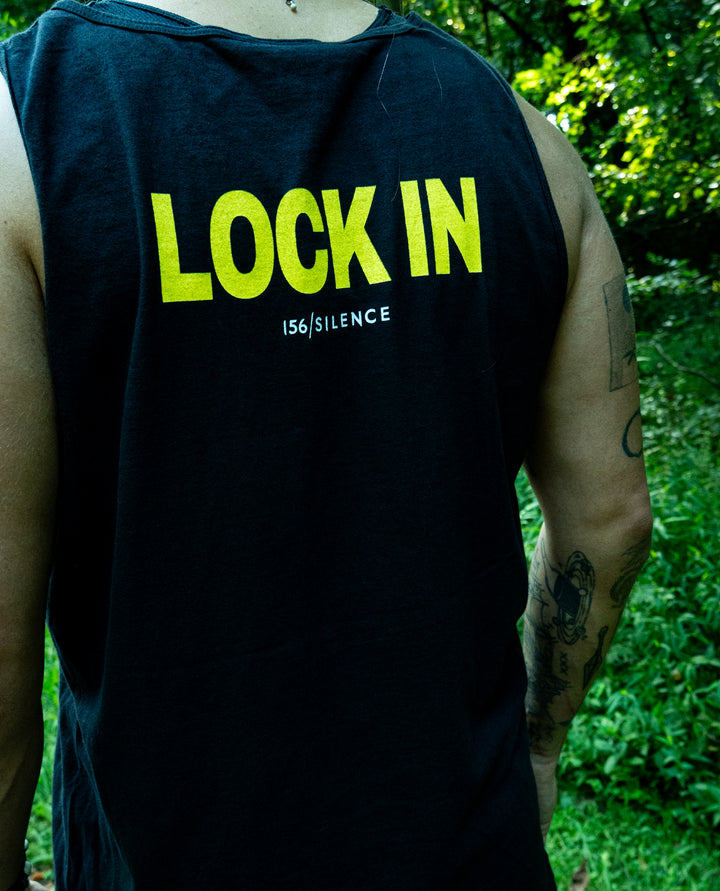 Person standing outdoors wearing a sleeveless black shirt, crafted with soft ring-spun cotton, featuring bold yellow text that reads LOCK IN and smaller white text below. Trees and greenery are in the background.