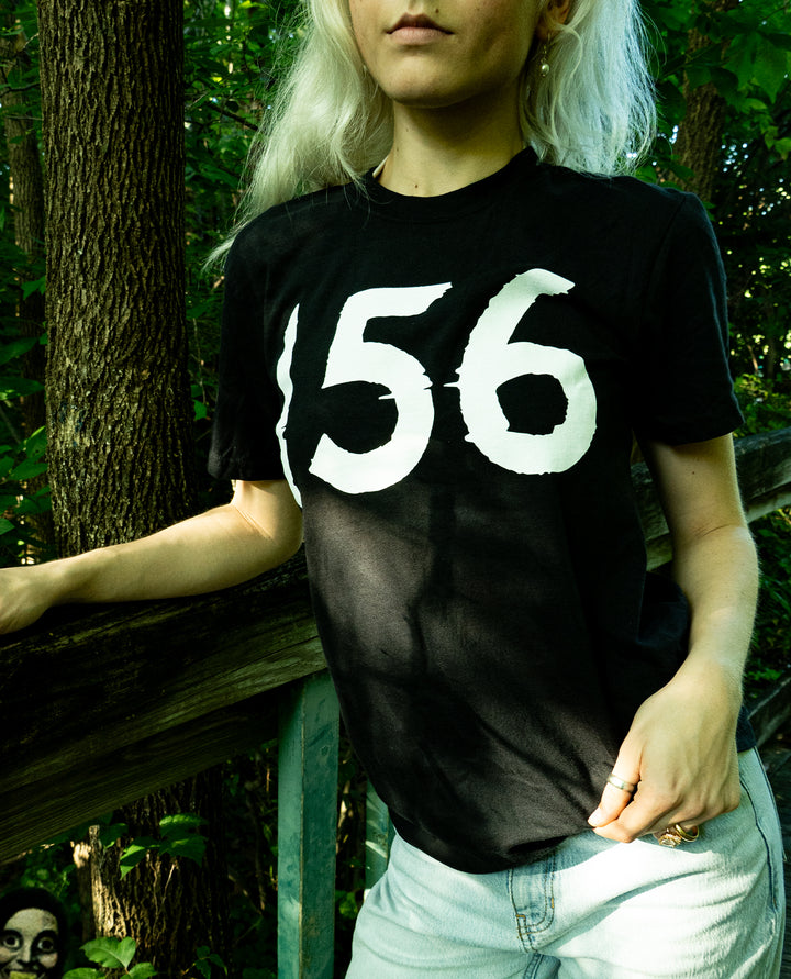 A person with long blonde hair wears a black heavyweight fleece t-shirt with the number 156 in large white print, standing in a sunlit, wooded area. A mysterious face is partially visible in the bottom left corner.