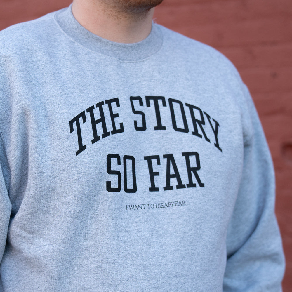 Someone wears a light gray sweatshirt from The Story So Far featuring bold black THE STORY SO FAR and smaller I WANT TO DISAPPEAR text, standing in front of a red brick wall.