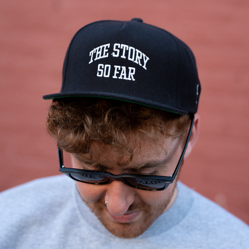 A person with curly brown hair wears a black embroidered The Story So Far snapback, sunglasses, and a grey shirt, looking down against a reddish wall.