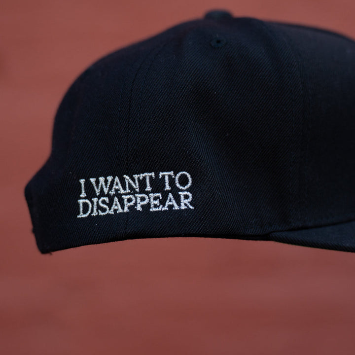 A black The Story So Far YP Classics 5 Panel Hat with white I WANT TO DISAPPEAR embroidery on the side, displayed against a blurred reddish-brown background.