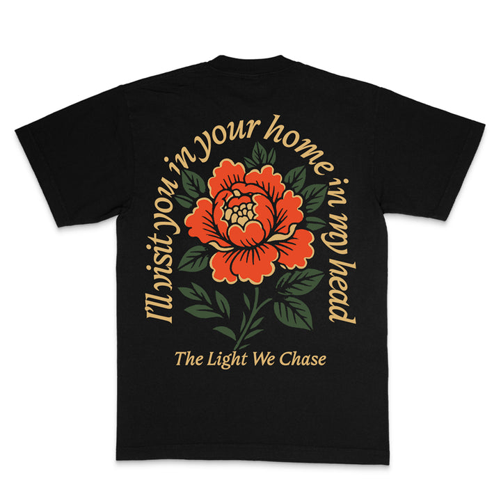 A Bloom Rose Black t-shirt features a large red and orange flower with green leaves on the back. Curved yellow text reads I’ll visit you; in your home, in my head above smaller text: The Light We Chase. Home Is In My Head inspired design.