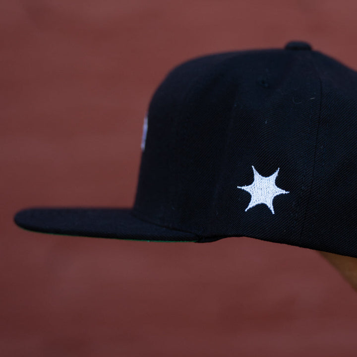 A black The Story So Far YP Classics 5 Panel Hat with a white star-shaped side design, displayed against a blurred reddish-brown background.