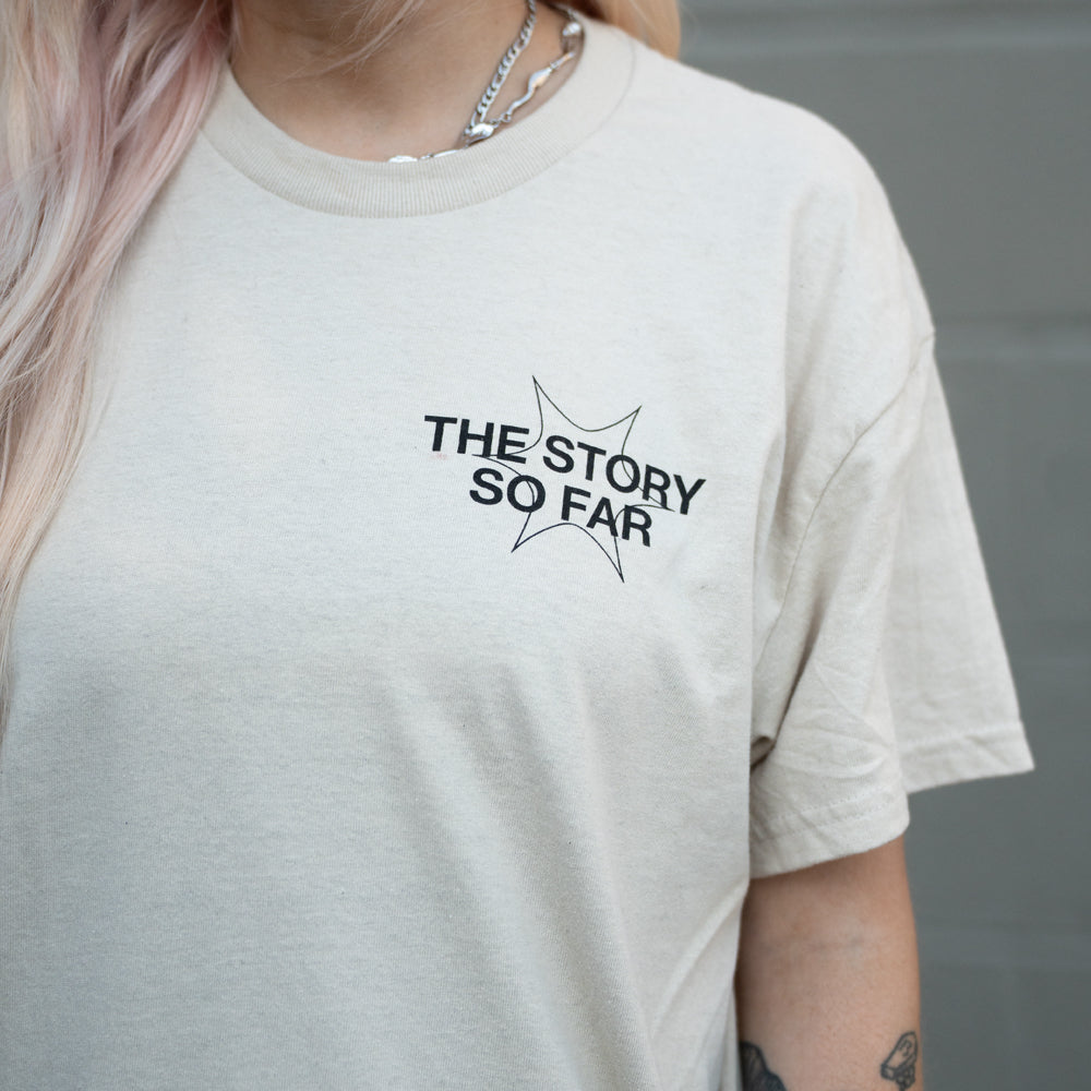 A person with long, light-colored hair wears a beige t-shirt by The Story So Far featuring a star graphic and band name on the chest, paired with a silver chain necklace.