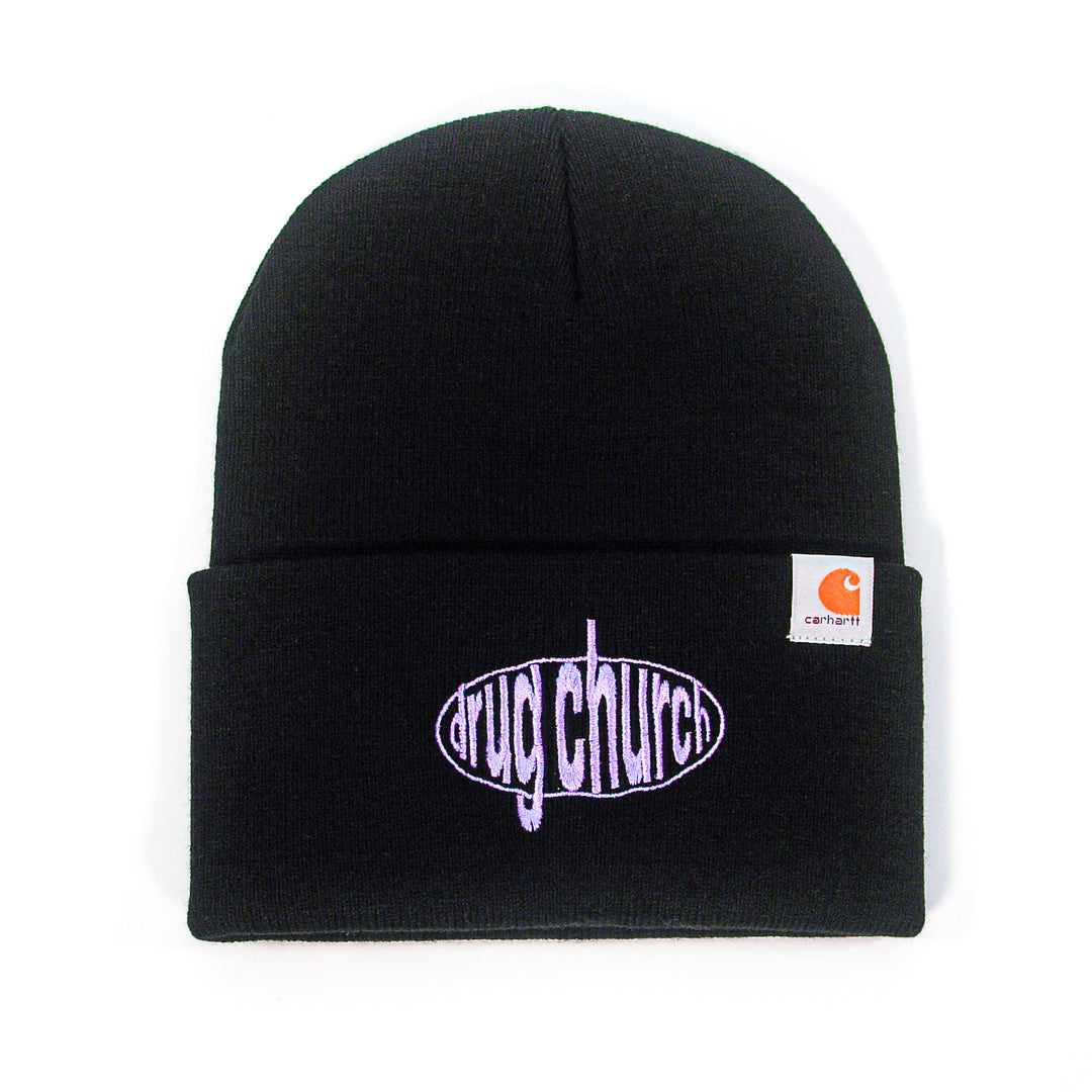 A black Drug Church beanie with a folded cuff features "drug church" in purple inside an oval on the front, plus a small Carhartt logo tag on the side—perfect for fans of embroidered beanies.