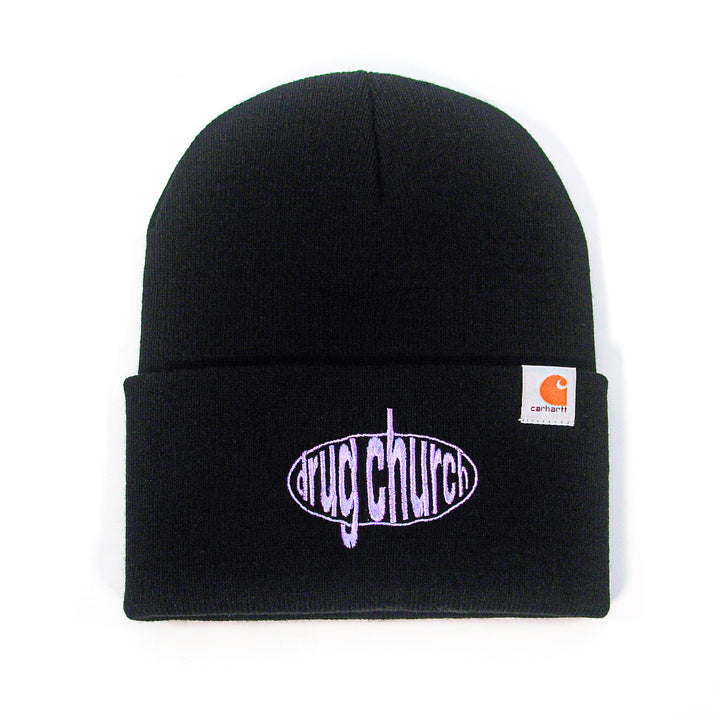 A black Drug Church beanie with a folded cuff features "drug church" in purple inside an oval on the front, plus a small Carhartt logo tag on the side—perfect for fans of embroidered beanies.