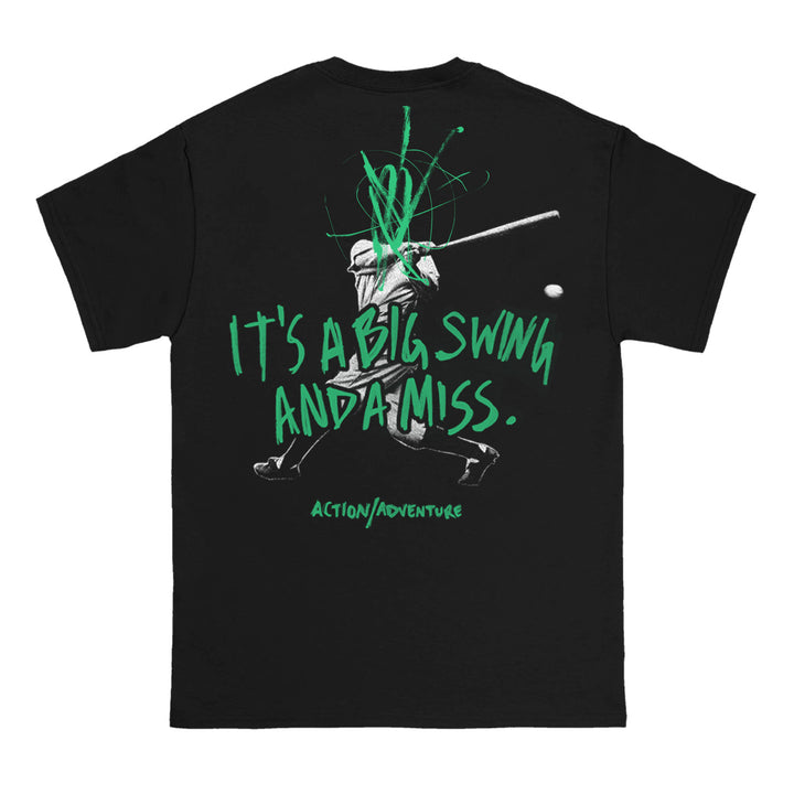 Black T-shirt by Action/Adventure featuring a grayscale baseball player swinging, with green paint marks and “IT’S A BIG SWING AND A MISS. ACTION/ADVENTURE.” Text; baseball flying past the batter.